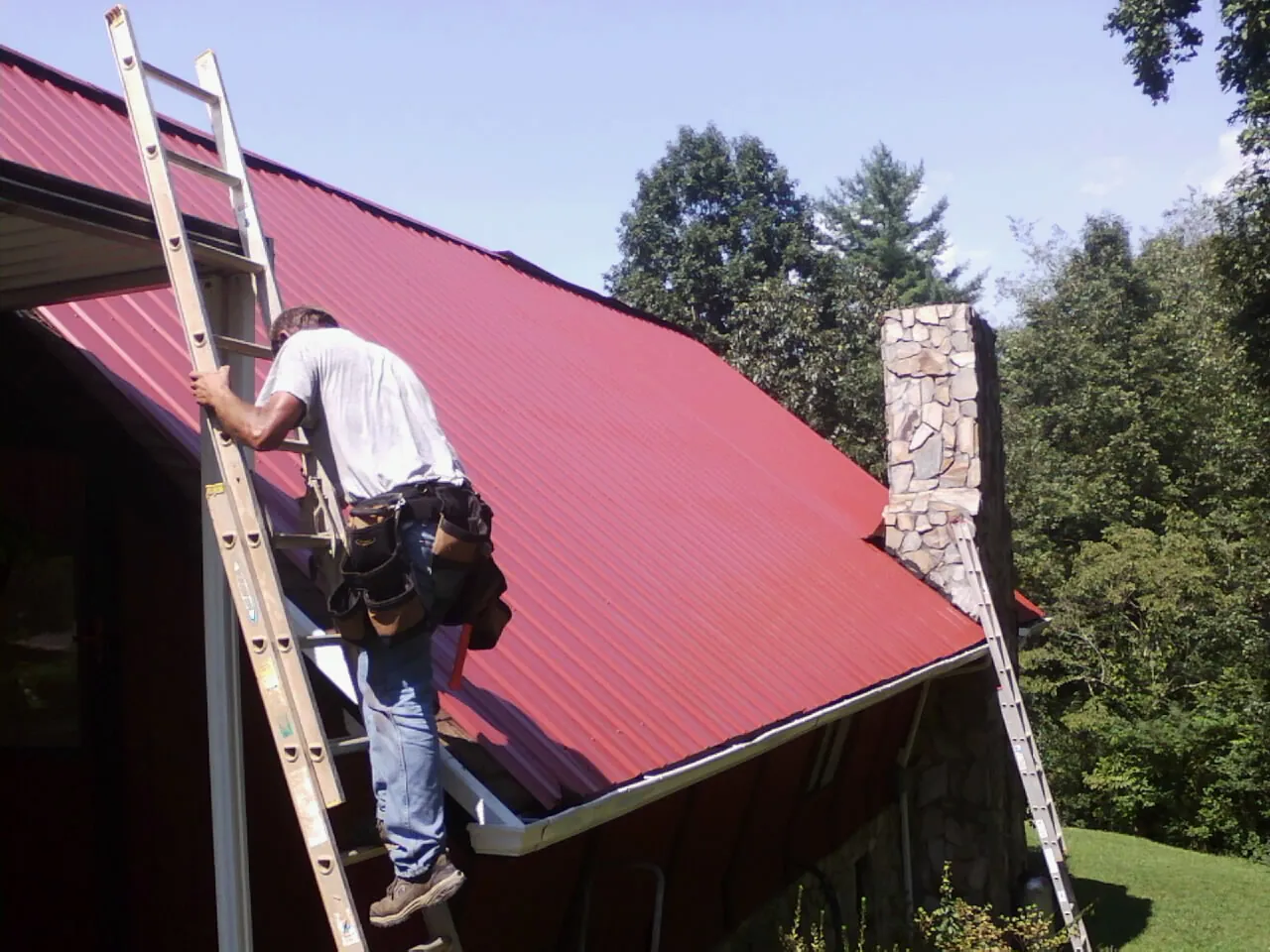 Roofer on a ladder beside a red metal roof and stone chimney — Soffit & Fascia Repair by Roofing Brothers in Sulphur